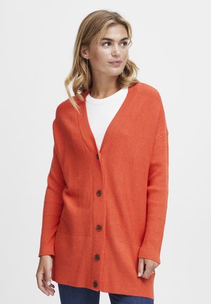 Cardigan orange côtelé avec un col en V, fermeture à boutons et manches longues. Texture douce et coupe décontractée, associé à un haut blanc et un jean.