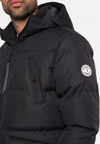 Veste matelassée noire avec capuche, fermeture éclair à l'avant, poche poitrine à boutons et écusson logo sur la manche. Texture lisse.