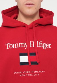 Red hoodie made of cotton blend, featuring "Tommy Hilfiger" embroidered in white, with a rectangular logo and established year below.