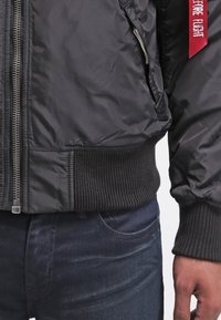 Gros plan d'une personne portant une veste noire zippée avec des poignets et un ourlet côtelés, un jean bleu foncé et une étiquette en tissu rouge sur la manche.