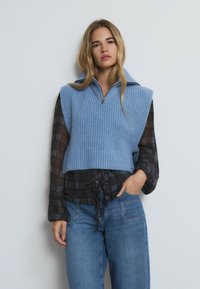 Sudadera sin mangas de punto en azul claro con textura acanalada, cremallera frontal y un cuello amplio, superpuesta a una camisa de manga larga a cuadros oscuros y jeans.