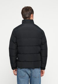 Uomo che indossa un giubbotto trapuntato nero e jeans blu, girato di spalle su uno sfondo bianco uniforme.