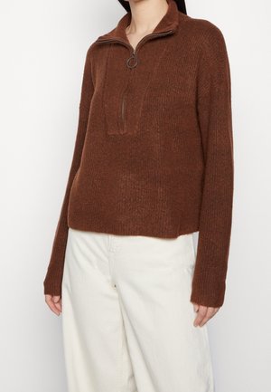 Vrouw draagt een bruin gebreide trui met lange mouwen, een halve ritssluiting en een ritslipje, gecombineerd met hoge witte broeken.