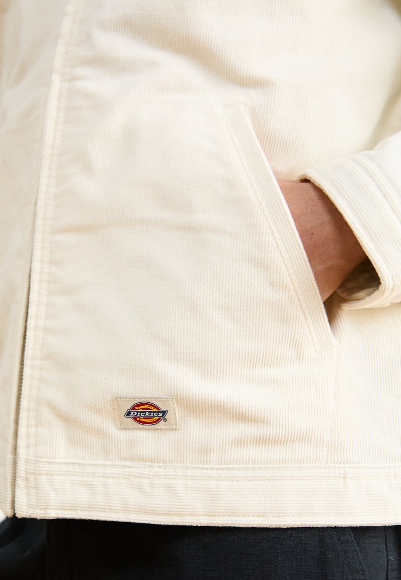 Veste en velours côtelé couleur crème avec une poche avant. Présente un patch logo Dickies à l'ourlet. Texture douce et motif en côtes.
