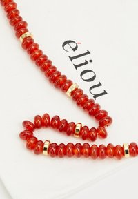 Collar de cuentas de vidrio rojo con acentos dorados, que presenta cuentas en forma circular y ovalada, exhibido sobre un paño blanco con el nombre de la marca "eliou."