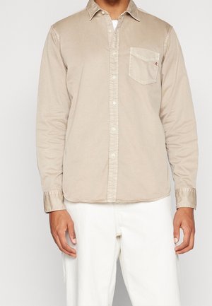 Uomo con una camicia beige a maniche lunghe con bottoni e taschino sul petto e pantaloni bianchi, in piedi davanti a uno sfondo semplice.
