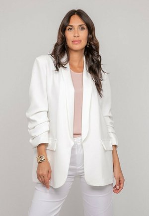 Femme aux longs cheveux foncés portant un blazer blanc et un pantalon blanc, un haut rose clair, des boucles d’oreilles dorées et un bracelet doré, debout devant un fond uni.