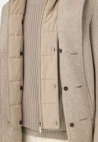 Béžový prošívaný kabát s pletenou texturou, vybavený vysokým límcem, zapínáním na knoflíky a jemnými akcenty. Nositelný přes světlý béžový svetr.