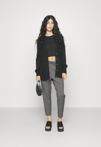 Svart cardigan med knapper, kort svart topp, grå mønstrede bukser og svarte platåsandaler. Liten svart håndveske og krøllete hår.