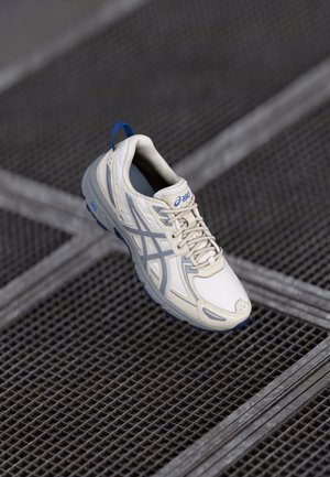 Chaussure de course Asics crème et bleue flottant au-dessus d'un sol en grille métallique avec des lignes diagonales et verticales.