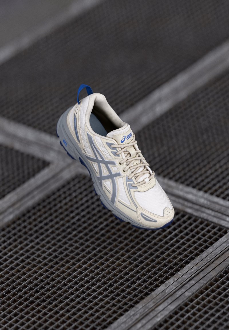 Chaussure de course Asics crème et bleue flottant au-dessus d'un sol en grille métallique avec des lignes diagonales et verticales.