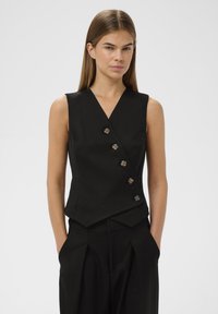 Črn brezrokavnik z V-izrezom, ki ima šest gumbov v kontrastni barvi, prilegajoč kroj in kotahem linijo. Gladka tekstura tkanine.