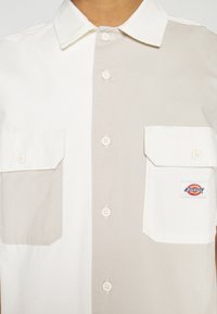 Camicia bicolore con pannelli bianchi e beige, due tasche sul petto e patch con logo. Caratterizzata da un colletto classico e chiusure con bottoni.