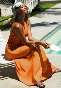 Robe maxi orange sans manches cintrée à la taille, assortie à des sandales noires avec des lanières décoratives. Un foulard léger et un éventail complètent le look.