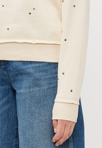 Gros plan d'une personne portant un jean bleu et un pull crème avec de petits clous argentés sur la manche et des poignets côtelés.