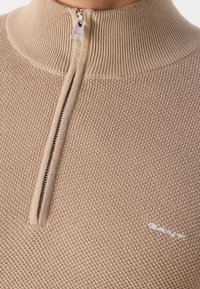 Maglione beige a maglia con colletto a zip a un quarto, zip argentata e piccolo logo bianco "GANT" ricamato sul petto.