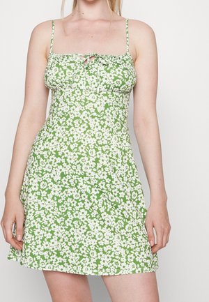 Frau trägt ein grünes Minikleid mit weißem Blumenmuster und dünnen Trägern, die an der Büste gebunden sind, steht vor einem schlichten Hintergrund.