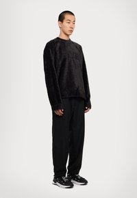 Μαύρος φλις μπλούζα με υφή, featuring ανάγλυφο λογότυπο, συνδυασμένη με μαύρο χαλαρό παντελόνι και μαύρα αθλητικά παπούτσια.