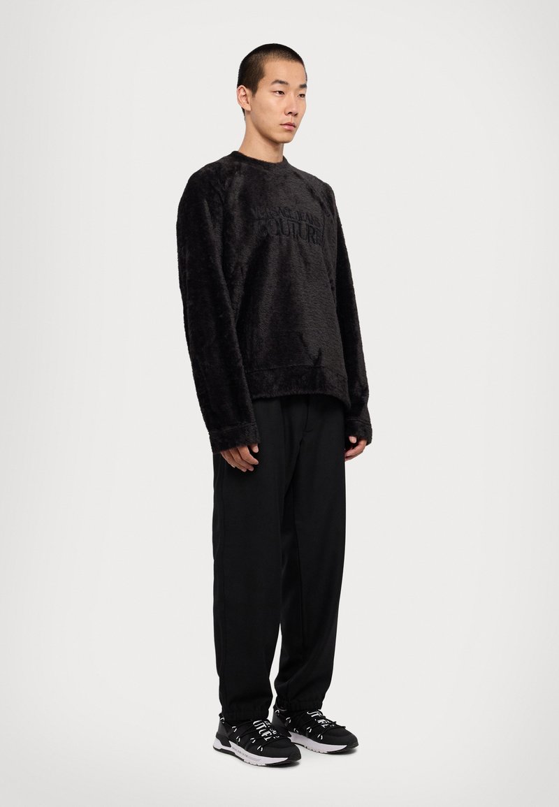 Μαύρος φλις μπλούζα με υφή, featuring ανάγλυφο λογότυπο, συνδυασμένη με μαύρο χαλαρό παντελόνι και μαύρα αθλητικά παπούτσια.