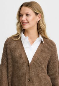 Brauner gestrickter Cardigan mit V-Ausschnitt, aus strukturiertem Stoff, zwei Knöpfen und lockeren Ärmeln. Wird über einem weißen Hemd mit Kragen getragen.