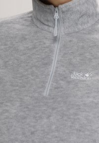 Szary polarowy sweter z wysokim kołnierzem, z ćwierć-zamkiem błyskawicznym oraz małym białym haftowanym logo po lewej stronie. Miękka tekstura.