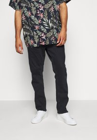 Kortärmad, mörkblommig skjorta med gröna och rosa blad på en marinblå bakgrund, i kombination med svarta byxor och vita sneakers.