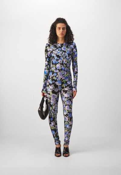 Long-sleeve, form-fitting jumpsuit in a black fabric with blue and green floral patterns. Paired with a black handbag and open-toe heels.