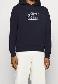Felpa con cappuccio navy in misto cotone, con logo frontale in testo grigio chiaro e polsini e orlo a coste. Indossata con pantaloni bianchi.