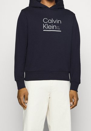 Sudadera con capucha navy hecha de una mezcla de algodón, que presenta un logotipo frontal en texto gris claro y puños y dobladillo acanalados. Combinada con pantalones blancos.