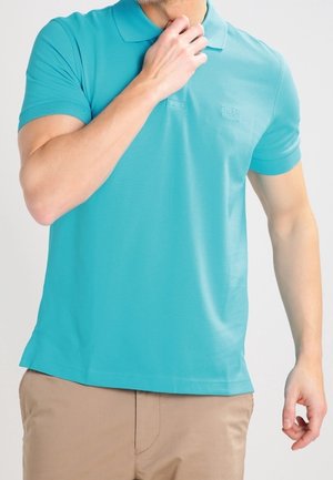 Homme portant un polo turquoise à manches courtes et un pantalon beige, la main touchant le menton, fond neutre.