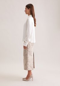 Hvid silke langærmet bluse sammen med en beige broderet nederdel med blomstermønstre og et sidesnit, suppleret med nude højhælede sko.