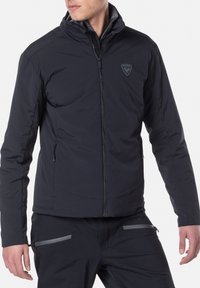 Zwarte geïsoleerde jas met een ritssluiting aan de voorkant, hoge kraag, twee ritssluitingen aan de zijkanten en een logo op de borst. Soepele stofstructuur.
