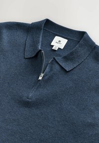 Tamsiai mėlynas zip polo marškinėliai, pasižymintys minkšta tekstūra, klasikinėmis apykaklėmis ir dalinio ilgio užtrauktuku priekyje. Prekės etiketė matoma viduje.