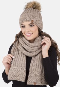 Bonnet en tricot beige avec pompon en fourrure et écharpe assortie. Les deux présentent des mailles texturées et une étiquette en cuir sur le bonnet.