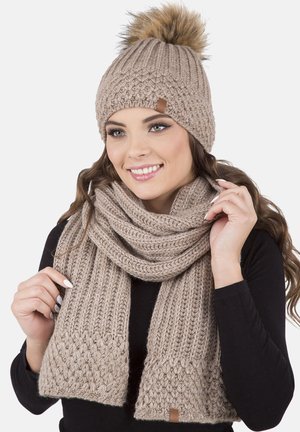 Berretto in maglia color cammello con pom-pom in pelliccia e sciarpa coordinata. Entrambi presentano punti di tessitura testurizzati e un'etichetta in pelle sul berretto.