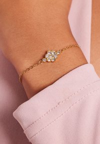 Gouden armband met een bloemontwerp, versierd met meerdere heldere edelstenen, bevestigd aan een delicatete ketting, gedragen om een pols naast een lichtroze stof.