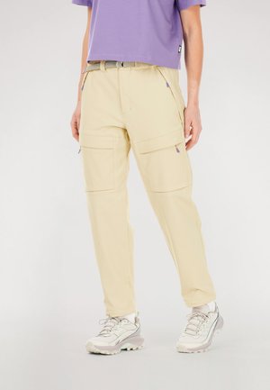 Light beige cargo pants feature two large front pockets, subtle texture, and adjustable drawstring waistband. Worn with a purple top and sneakers.