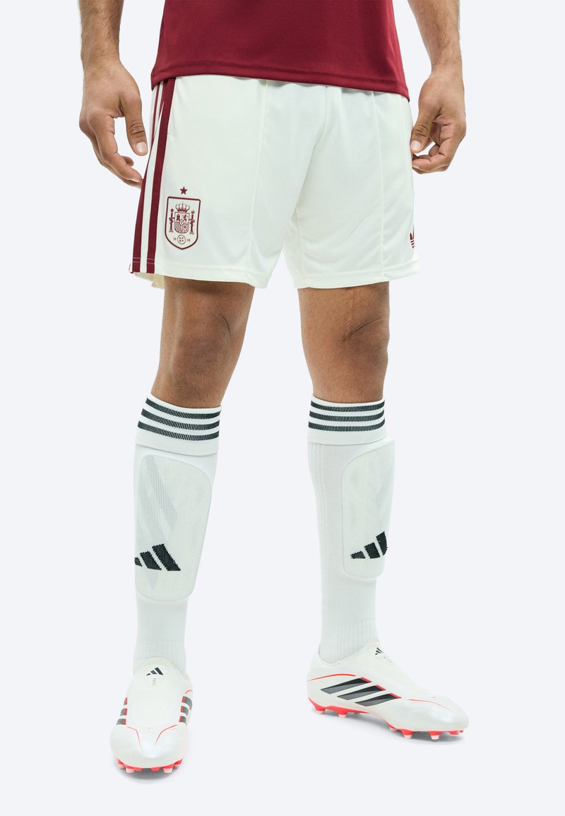 Hommes portant des shorts de football blancs avec le blason de l'Espagne, des chaussettes blanches à rayures noires, des crampons blancs et des protège-tibias, debout devant un fond blanc.