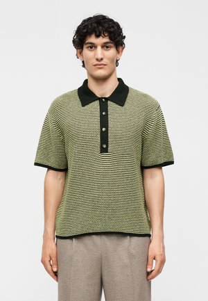 Νέος άνδρας με σγουρά μαλλιά που φοράει κοντομάνικο πικέ πόλο με μαύρο γιακά και κουμπιά, σε συνδυασμό με μπεζ παντελόνι.