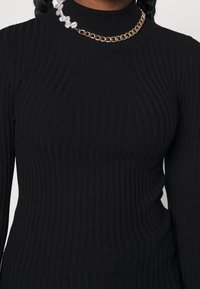 Svart ribbad polotröja med långa ärmar, prydd med ett strukturerat stickat mönster. Accentuerad med ett lager av guld- och pärlhalsband.