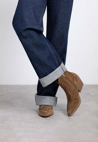 Stivaletti in camoscio marrone con perforazioni decorative, indossati con jeans di denim scuri arrotolati e con risvolti grigi chiari.