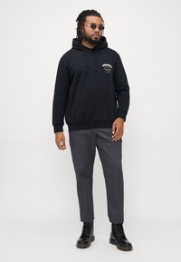 Black hoodie with front pocket and logo, paired with gray cargo pants. Finished with black lace-up boots and round sunglasses.