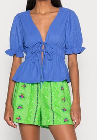 Blue ribbed top with puff sleeves and a deep V-neck, tied at the waist. Paired with green shorts featuring floral patterns and accents.