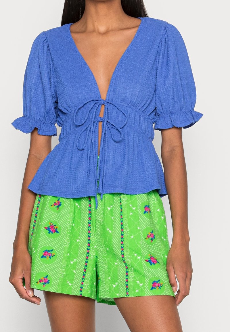 Blue ribbed top with puff sleeves and a deep V-neck, tied at the waist. Paired with green shorts featuring floral patterns and accents.