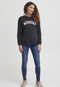 Felpa nera con la scritta "NEWPORT" in lettere bianche, abbinata a jeans skinny blu e sneakers beige. Il modello si trova su uno sfondo neutro.