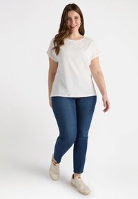 Top blanco de manga corta con detalles bordados en el cuello, combinado con jeans ajustados de color azul oscuro y zapatillas claras, sobre un fondo sencillo.