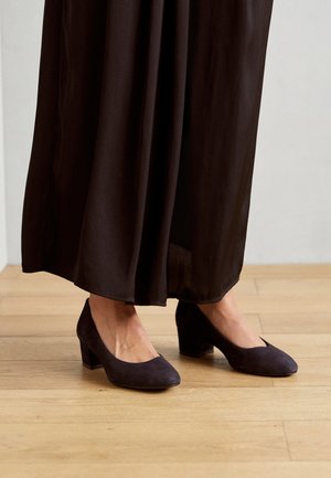 Des chaussures à talons blocs en daim violet foncé avec un bout rond, positionnées sur un sol en bois, partiellement recouvertes par une jupe noire fluide.