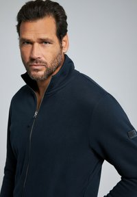 Chaqueta de forro polar azul marino con cremallera y cuello alto, que cuenta con un parche en la manga izquierda y una textura suave. Cremallera visible en la parte delantera.