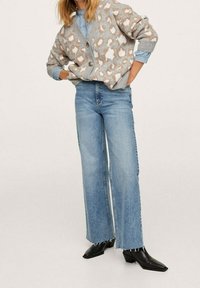Cardigan grigio con macchie leopardate bianche e marroni, chiusura con bottoni, abbinato a jeans chiari a vita alta e stivali neri con punta a punta.