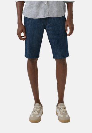Mann trägt dunkelblaue Jeansshorts, hellbeige Sneaker und ein hellgraues Hemd mit Knöpfen, steht vor einem schlichten Hintergrund.